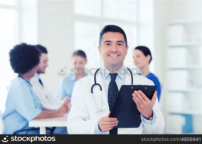 clinic, profession, people and medicine concept - happy male doctor with clipboard over group of medics meeting at hospital