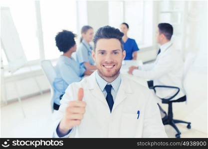 clinic, profession, people, gesture and medicine concept - happy male doctor over group of medics meeting at hospital showing thumbs up gesture. happy doctor over group of medics at hospital