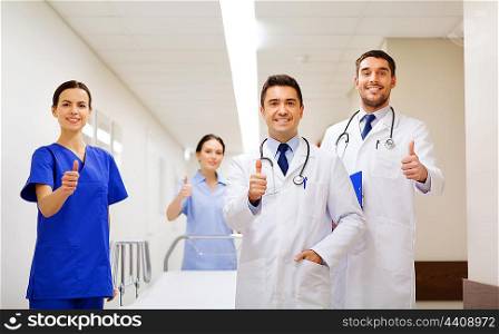 clinic, profession, people, health care and medicine concept - group of happy medics or doctors with gurney showing thumbs up at hospital corridor