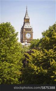 Clock face on the famous landmark clock tower known as Big Ben in London, England visible through the trees of a London park.