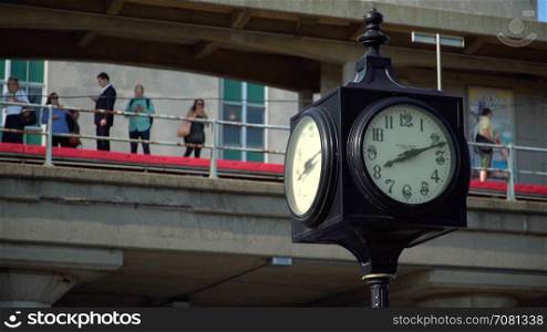 Clock in foreground with commuters waiting on a train station