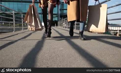Cloesup of sexy elegant female legs in high heels walking on pedestrian bridge at sunset. Two beautiful women carrying shopping bags strilling on bridge over shopping mall background. Slow motion.
