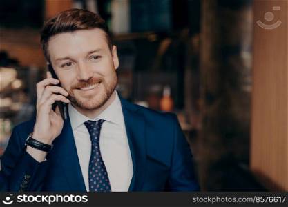 Close portrait of good looking entrepreneur with beard and hairdo in formal suit talking on cellphone about successfully completed financial project, looking aside and smiling while standing in cafe. Close portrait of good looking happy financial entrepreneur enjoying talk on phone