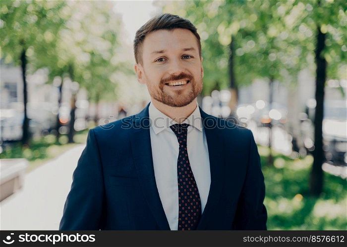 Close portrait of smiling handsome young bearded man in dark blue suit walking outside in sunny day along green alley in city centre, happy to receive promotion at work today. Close portrait of smiling handsome young bearded businessman outdoors