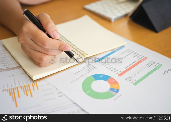 Close shot of businesswoman hands holding a pen writing something on the paper on the foregroundin office. Recording concept. Using smartphone tablet. Freelance Work, Business concept