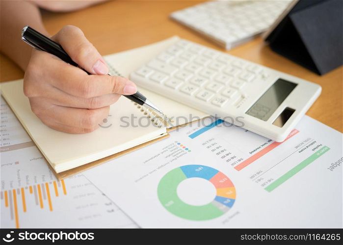 Close shot of businesswoman hands holding a pen writing something on the paper on the foregroundin office. Recording concept. Using smartphone tablet. Freelance Work, Business concept