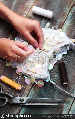 Close shot of female hands decorating purse with sequins using thread needle and scissors following diy ideas on wooden table
