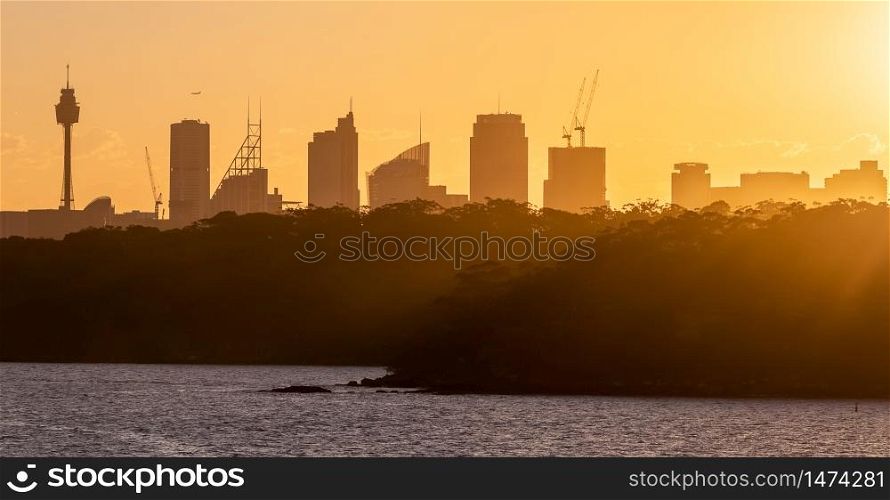 Close silhouette view of Sydney downtown with trees in the foreground. Sun rays spreading an amazing orange color cast over the trees. Beautiful orange sky in the background