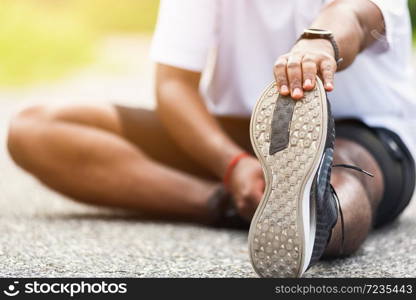 Close up Asian young athlete sport runner black man wear watch he sitting pull toe feet stretching legs and knee before running at outdoor street health park, healthy exercise before workout concept