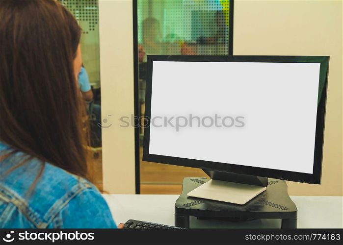 Close up back view of a young businesswoman looking at a screen while working on a desktop computer at her desk at the office. Modern and comfortable interior office. Mockup blank white screen with copy space.