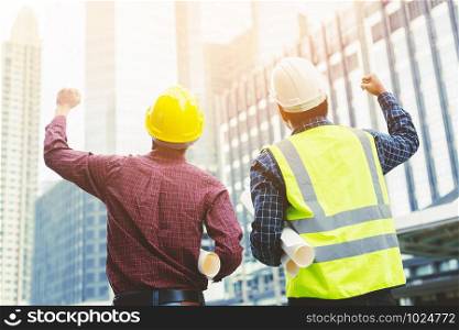 Close up back view of engineering male two construction worker wear safety helmet holding roll paper drawing model plan project. engineer standing raised arms fist cheerful show project success.