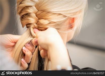 Close up back view of female hands braiding a pigtail to young blond woman at beauty salon. Female hands braiding pigtail to woman