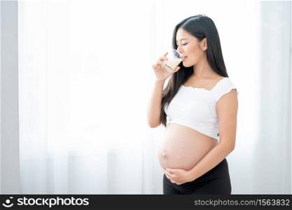 Close up beautiful Asian pregnant woman is drinking milk and stand in front of white curtain with morning light. Concept of good healthy food for mother and support baby in belly of people.