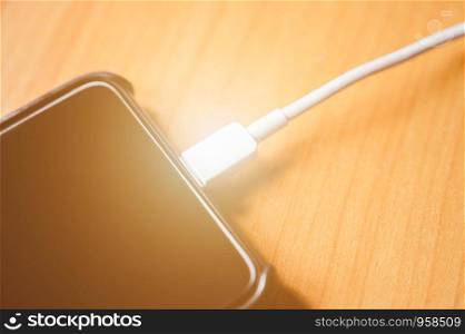 Close-up black color mobile smart phone charging on wooden table near the window.