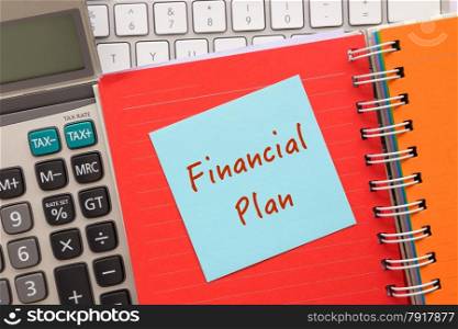 "Close up blue notepad with words " Financial Plan " putting on on colorful paper book with keyboard and calculator on background"