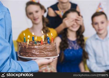 close up boy s hand bringing chocolate cake birthday girl with covered eyes by her friend