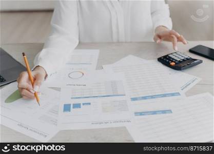 Close-up businesswoman calculating expenses, managing project budget, sitting at desk with documents. Woman counts finances, preparing report in charts and graphs, analyzing financial statistics.. Female business person works calculating project budget, analyzing financial statistics, close-up