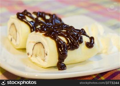 close up crepe rolls filled with white cream and banana, topping with chocolate syrup served with whipping cream on white dish