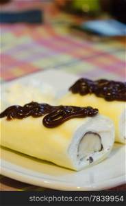 close up crepe rolls filled with white cream and banana, topping with chocolate syrup , served on white dish