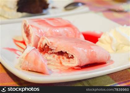 close up crepe rolls filled with white cream and strawberry, topping with strawberry syrup served with whipping cream on dish