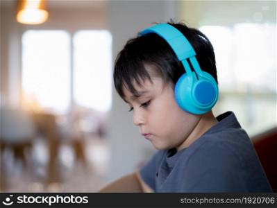 Close up face Happy kid wearing headphones listening to music while drawing with blurry living room background, Indoors side view portrait Cute child boy enjoy creative activity at home on weekend.
