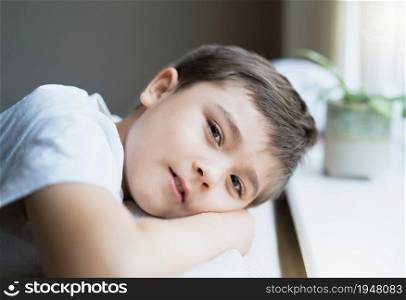 Close up face of happy young boy lying head on hands relaxing at home, Candid shot kid looking at camera with smiling face sitting alone next to window, Positive children for Children day concept