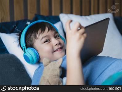Close up face young boy wear wireless headphone listening music lying in bed,Happy Kid playing game on tablet, Smiling child in blue pajamas relaxing in bed room in morning before go to school