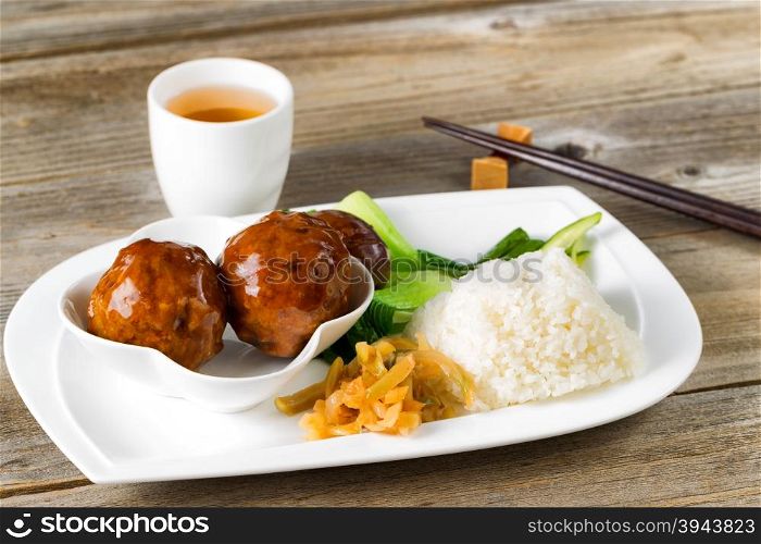 Close up front view Asian saucy meatballs, rice and bok choy on white plate. Chopsticks and green tea in background.
