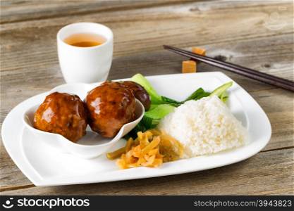 Close up front view Asian saucy meatballs, rice and bok choy on white plate. Chopsticks and green tea in background.