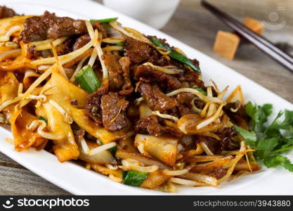 Close up front view of freshly cooked spicy beef, bean sprouts, onion, and tofu with parsley as garnish and chopsticks in holder on rustic wood.