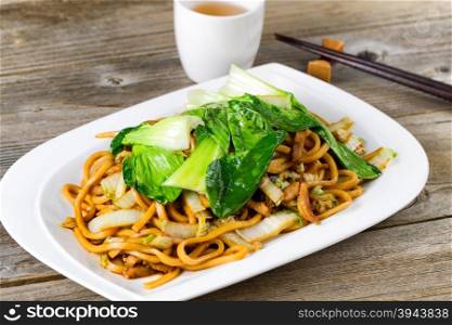 Close up front view of spicy cooked noodles, onion, bok choy, and chicken slices. Chopsticks and tea in background on rustic wood. Selective focus on middle of dish.