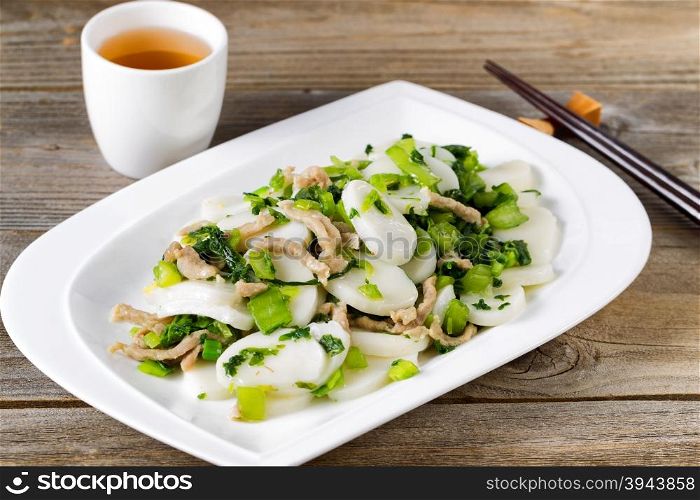 Close up front view of stir fry consisting of sliced sticky rice cakes, onion, and chicken. Chopsticks and tea in background on rustic wood. Selective focus on front of dish.