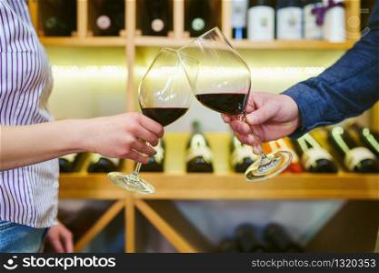 Close up front view on male and female caucasian hands holding glasses of red wine toasting in front of the shelf man and woman celebrating concept