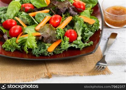 Close up front view, selective focus on lower right corner of plate, of fresh green salad with carrots, cherry tomato, basil, baby kale, and lettuce on rustic white wooden table.