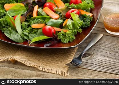 Close up front view, selective focus on lower right corner of plate, of fresh green salad with carrots, cherry tomato, basil, baby kale, and lettuce on rustic wood.