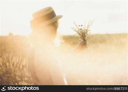 Close up girl in straw hat with bouquet surrounded by sunlight glare concept photo. Side view photography with meadow on background. High quality picture for wallpaper, travel blog, magazine, article. Close up girl in straw hat with bouquet surrounded by sunlight glare concept photo