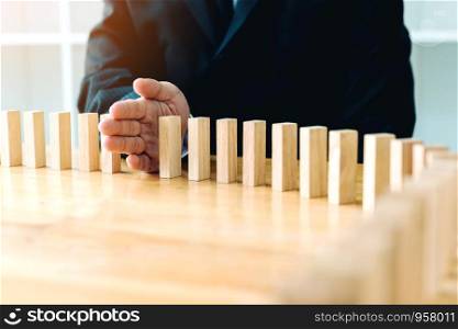 Close up hand businessman stopping wooden block from falling in the line of domino with risk concept.
