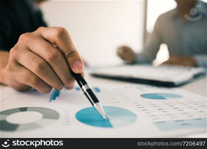 Close up hand of a businessman holding a pen pointing to graph of financial statements or company profits and analyzing on the desk at the office room.