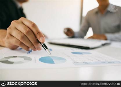 Close up hand of a businessman holding a pen pointing to graph of financial statements or company profits and analyzing on the desk at the office room.