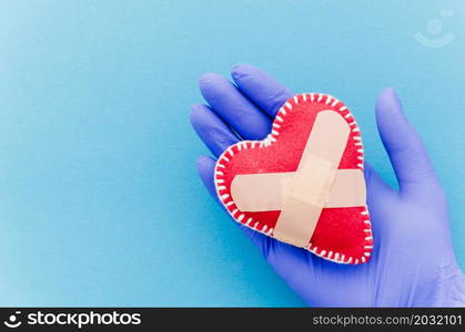 close up hand wearing surgical gloves holding heart shaped stitched textile heart with crossed bandages blue backdrop