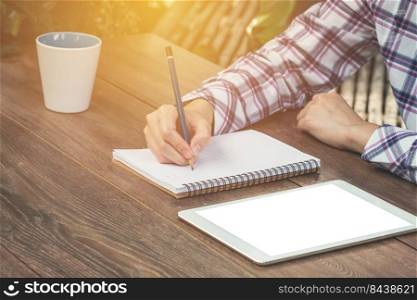Close up hand woman writing notebook and blank tablet on wood table with sunlight. Vintage toned.