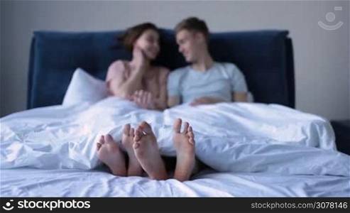Close-up human male and female feet under the white blanket with out of focus laughing couple in background. Couple&acute;s feet under the blanket in modern styled bed in the morning.