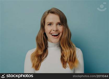 Close-up image of attractive happy young lady in casual clothes with wavy light-brown hair looking at camera with beaming smile laughing over funny joke or anecdote isolated on blue studio background. Attractive happy young lady looks at camera with beaming smile laughing over funny joke or anecdote