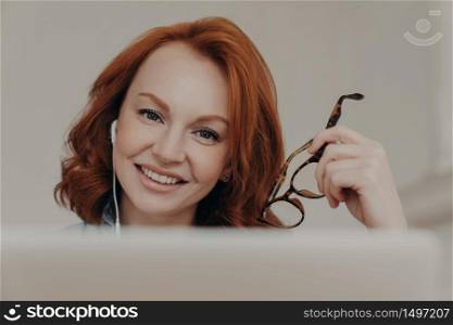 Close up image of ginger positive European woman smiles gently, enjoys online communication, watches tutorial webinar, gets new skills, sits in front of laptop computer, holds transparent glasses