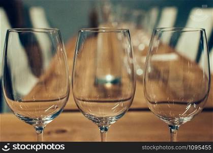 Close up image of three empty glasses on a wooden table, served for wine tasting event. White chairs in the background. Bar or restaurant interior, subdued light. Selective focus, film grain effect