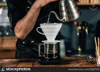 Close up image of young male barista pouring boiling water from kettle to drip coffee maker on wooden table. Barista wearing dark uniform.