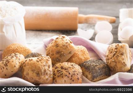 Close Up image with freshly baked buns, decorated with poppy seeds, sesame, flax and sunflower seeds, with flour, eggs and a rolling pin in the background.