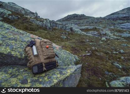 Close up large rucksack on rock at highland concept photo. Front view photography with mountains under cloudy sky on background. High quality picture for wallpaper, travel blog, magazine, article. Close up large rucksack on rock at highland concept photo