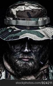 Close up, low key portrait of bearded commando fighter, army special forces soldier, private military company mercenary in boonie hat, tactical radio headset, black and green face camouflage paint. Commando soldier in boonie hat close up portrait