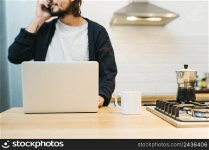 close up man talking mobile phone with laptop white mug kitchen counter